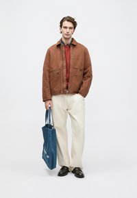 Jachetă maro cu fermoar frontal și buzunar pe piept, cămașă roșie dedesubt, pantaloni crem și o geantă de umăr albastră. Pantofi negri completează ținuta.