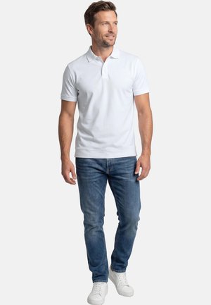 Man draagt een wit poloshirt met korte mouwen, blauwe spijkerbroek en witte sneakers, staand tegen een effen lichte achtergrond.