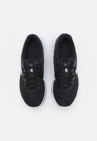 Chaussures de sport noires avec tige en mesh, design à lacets, semelle blanche et accent de marque sur la languette. Détails texturés partout.