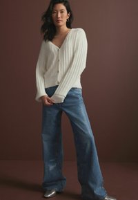 Vit, ribbad stickad cardigan med V-ringning och knappar, tillsammans med blå denimbyxor med vida ben och prickigt mönster. Silverfärgade spetsiga skor.