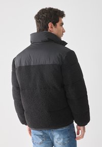 Uomo che indossa una giacca in pile nera con spalle imbottite e jeans azzurri, rivolto di lato su uno sfondo bianco semplice.