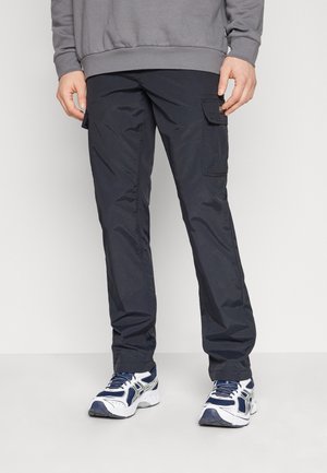 Mężczyzna w czarnych spodniach typu cargo z bocznymi kieszeniami, szarej bluzie i biało-niebieskich butach do biegania, stojący na jednolitym tle.