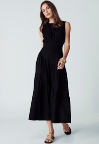 Črna brezrokavna obleka z oprijeto korzetno zgornjim delom in nagubanih naborkom. Ima gladko teksturo in geometrijske oblikovalske elemente.