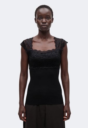 Femme portant un haut noir sans manches avec des détails en dentelle sur la poitrine et les épaules, associé à un pantalon noir ample, debout devant un fond uni.