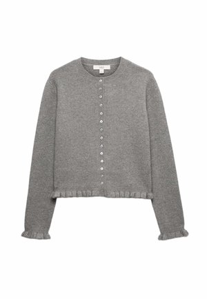 Donna con capelli lunghi e mossi che indossa un maglione grigio e una gonna, tenendo una borsa nera dietro la schiena su uno sfondo uniforme.