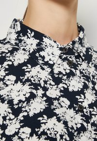 Chemise bleu marine avec motif floral blanc, col pointu et trois boutons noirs visibles, portée par une personne sur un fond neutre.