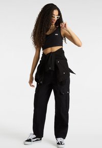 Zwart crop top met Vans-logo, zwarte cargo broeken met drukknopzakken, zwarte spijkerjasje om de taille geknoopt, witte sneakers met zwarte accenten.