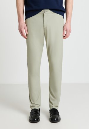 Uomo con pantaloni beige aderenti, mocassini neri, camicia blu scuro, in piedi davanti a uno sfondo chiaro e uniforme.