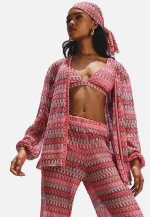 Drei-Teile-Outfit in Pink mit Muster, bestehend aus einem Bralette, einer lockeren Jacke und einer enganliegenden Hose. Der Stoff hat wellenförmige Linien in kontrastierenden Farben und eine strukturierte Optik.