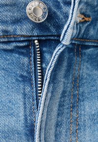 Gros plan sur la fermeture éclair d’un jean en denim bleu partiellement ouverte avec un bouton en métal visible et des coutures orange.
