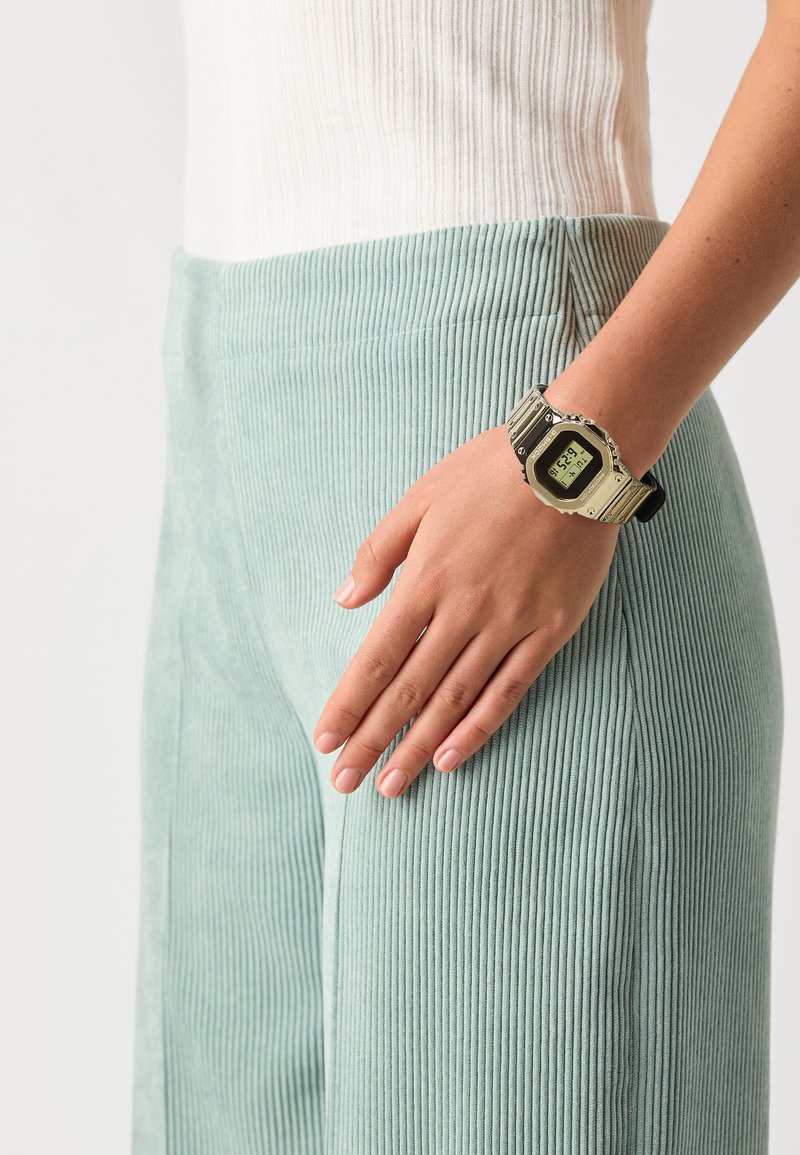 Orologio digitale con cinturino nero e hardware argentato; il display mostra l'ora. La mano poggia su pantaloni a coste verde menta. Top a coste bianco visibile.