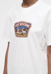 Λευκό T-shirt με γραφικό δύο κουνελιών και καρότων σε ένα τοπίο, με το κείμενο "LOVE MOSCHINO" πάνω από αυτό.