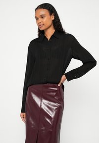 Melna, garām piedurknēm krekls ar vertikālām krokojumiem un pogu aizdari, kopā ar spīdīgu, bordo ādas svārku ar sānu šķēlumu.