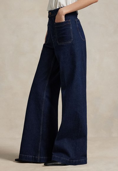 Asmuo dėvi tamsiai mėlynas, plačiomis kojomis džinsus su lopinėtomis kišenėmis priekyje ir gale, derinančias su juodais smailiais batais ir įkišta balta palaidine.