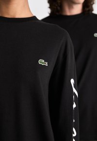 Schwarzes Langarm-T-Shirt mit einem kleinen grünen gestickten Krokodil-Logo auf der Brust und einem weißen Textdesign auf dem Ärmel.