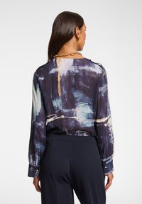 Blusa a maniche lunghe con una stampa astratta blu e viola, caratterizzata da dettagli dorati sui polsini e un design ampio e fluido.