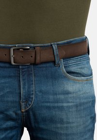 Cinturón trenzado marrón con una hebilla de metal rectangular, usado sobre unos pantalones vaqueros de mezclilla azules. Los vaqueros tienen costuras en contraste y un corte clásico.
