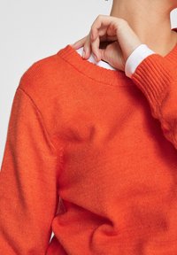Pull orange vif avec un col et des poignets côtelés, confectionné dans un tissu à la texture douce. Une chemise blanche est visible sous le col.