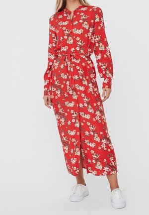 Femme portant une robe midi rouge à fleurs à manches longues avec ceinture à la taille, associée à des baskets blanches, debout devant un fond uni.