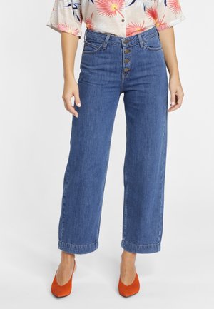 Vrouw draagt blauw wijde high-waist jeans met vier knopen aan de voorkant, een gebloemde blouse en oranje hoge hakken, staand.