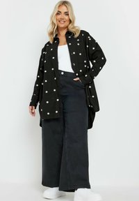 Camicia nera con bottoni, ricamata con margherite bianche, abbinata a pantaloni neri a gamba larga e sneakers bianche. Tessuto liscio, design informale.