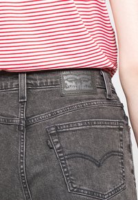 Jeans de mezclilla gris oscuro de Levi's con una etiqueta de cuero negro, que presentan dos bolsillos traseros y un detallado de costura distintivo, parcialmente cubiertos por una camisa a rayas rojas y blancas.