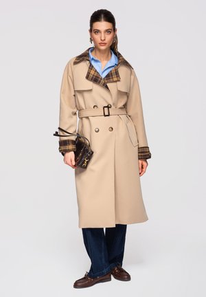 Donna che indossa un trench beige con cintura, colletto e polsini a quadri, camicia azzurra, jeans scuri, mocassini marroni, con una piccola borsa marrone in mano.