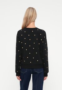 Sweatshirt noir avec des motifs de cœurs dorés, poignets et ourlet côtelés. Texture lisse et encolure ras du cou. Porté avec un jean bleu.