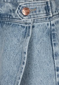 Lichtblauwe denimstof met een vervaagde textuur, voorzien van een koperen knop met het woord "BIZUU" in reliëf, met zichtbare gestikte naden.