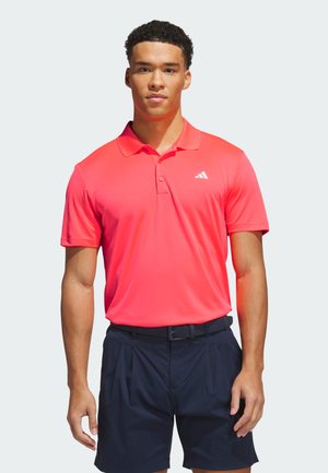 Muškarac obučen u svijetlo koraljasti Adidas polo i crne kratke hlače, stoji s opuštenim rukama na svijetloj pozadini.