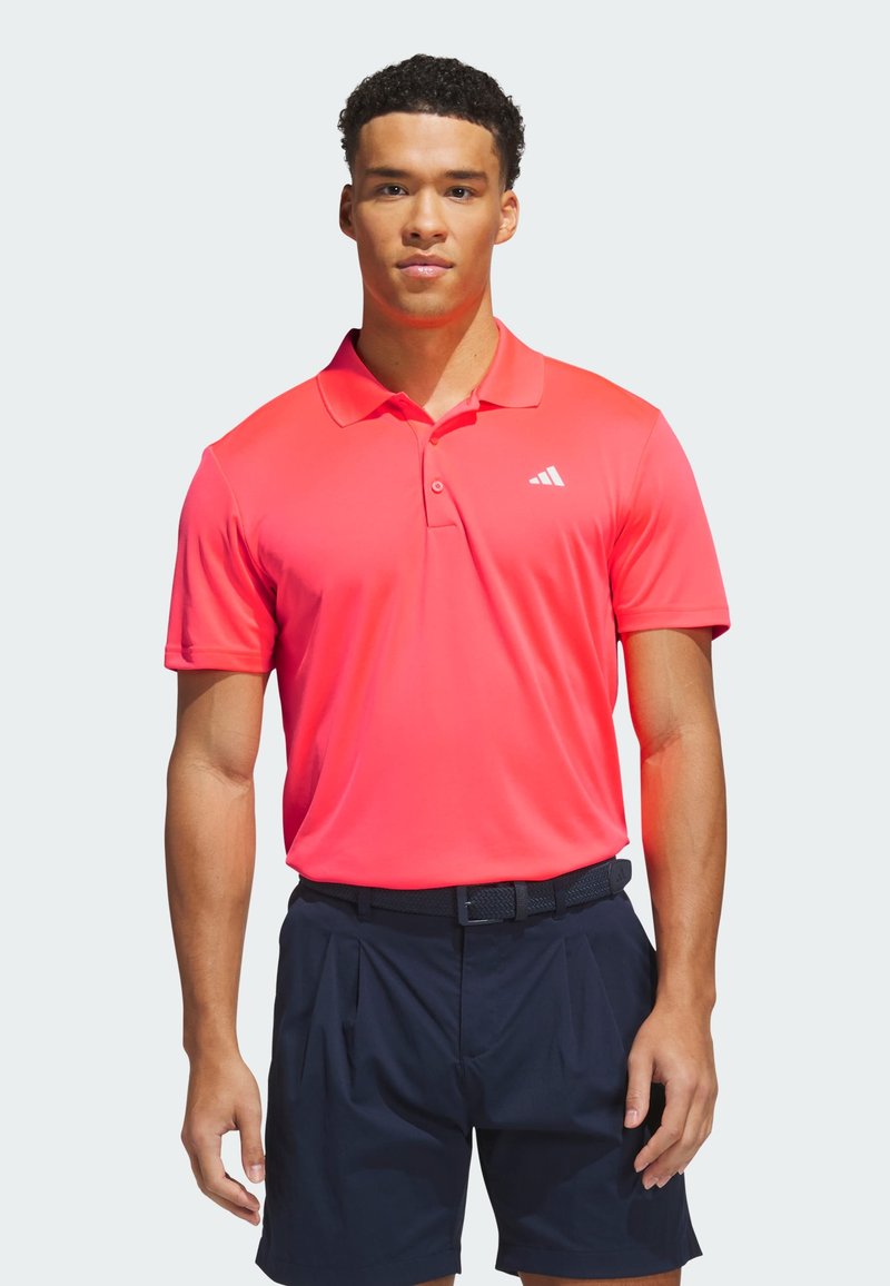 Homme portant un polo Adidas corail vif et un short noir, debout avec les bras détendus devant un fond clair.
