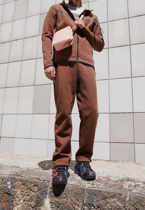 Mężczyzna ubrany w brązowy dres, czarno-czerwono-niebieskie trampki oraz różową torbę Nike na pasku, stojący na kamiennej krawędzi na tle ściany z płytek.