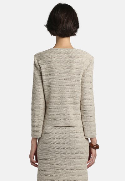 Femeie cu păr scurt și închis la culoare, purtând un top bej cu mâneci lungi și o fustă asortată, stând cu spatele spre privitor.