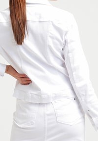 Giacca di denim bianco con design aderente, colletto a camicia e tasche posteriori. Presenta cuciture a vista e un logo sul bordo.