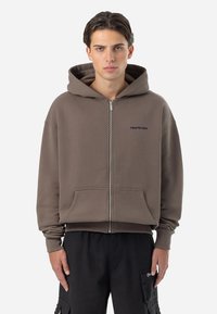 Sudadera con cremallera marrón de textura suave, con una gran capucha y bolsillos frontales. Presenta un logo sutil en el pecho. Fabricada en una mezcla de algodón.