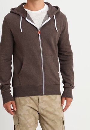 Homme portant un sweat à capuche marron zippé avec des cordons blancs et un pantalon cargo beige à motif camouflage, debout devant un fond uni.