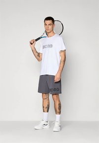 Valge T-särk kirjaga "BORG", hallid lühikesed püksid küljeribaga, seisab tennise reketiga, kannab valgeid tossusid ja sokke.