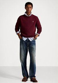 Sweatshirt borgonha, camisa de botão azul às riscas por baixo, jeans azul desbotados e sapatos castanhos com atacadores. Look casual em camadas.