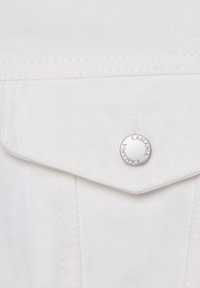 Valge teksariie lähivõte, mis näitab taskuklapiga topelttikkimist ja ümarat hõbedast pressnööpi, millele on graveeritud "LAMANIA".