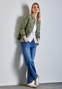 Licht olijfgroene bomberjack met een textuurafwerking, witte overhemden, blauwe brede spijkerbroek en witte sneakers met beige accenten.