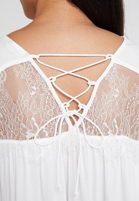 Abito bianco con schiena in pizzo e lacci incrociati. Tessuto morbido e fluttuante, decorato con motivi floreali e dettagli trasparenti.