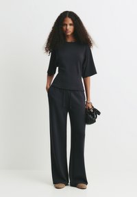 Femme aux cheveux bouclés portant un haut noir ample et un pantalon large, tenant un sac à main noir, se tenant contre un fond blanc.