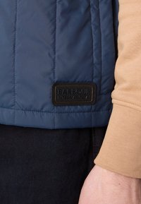 Casaco acolchoado azul-marinho com um rótulo preto da Barbour International, usado sobre uma camisa de manga longa bege, combinado com calças escuras.