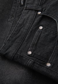 Primo piano della tasca di una giacca di denim nera con rivetti metallici e cuciture visibili su tessuto strutturato.