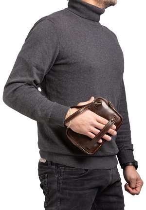 Borsa in pelle marrone con cerniera e tracolla, tenuta in mano. La persona indossa un maglione a collo alto grigio scuro e jeans neri.