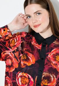 Langærmet skjorte i gennemsigtigt stof med blomsterprint i rød og orange, med en sort krave og sorte knapper langs fronten.