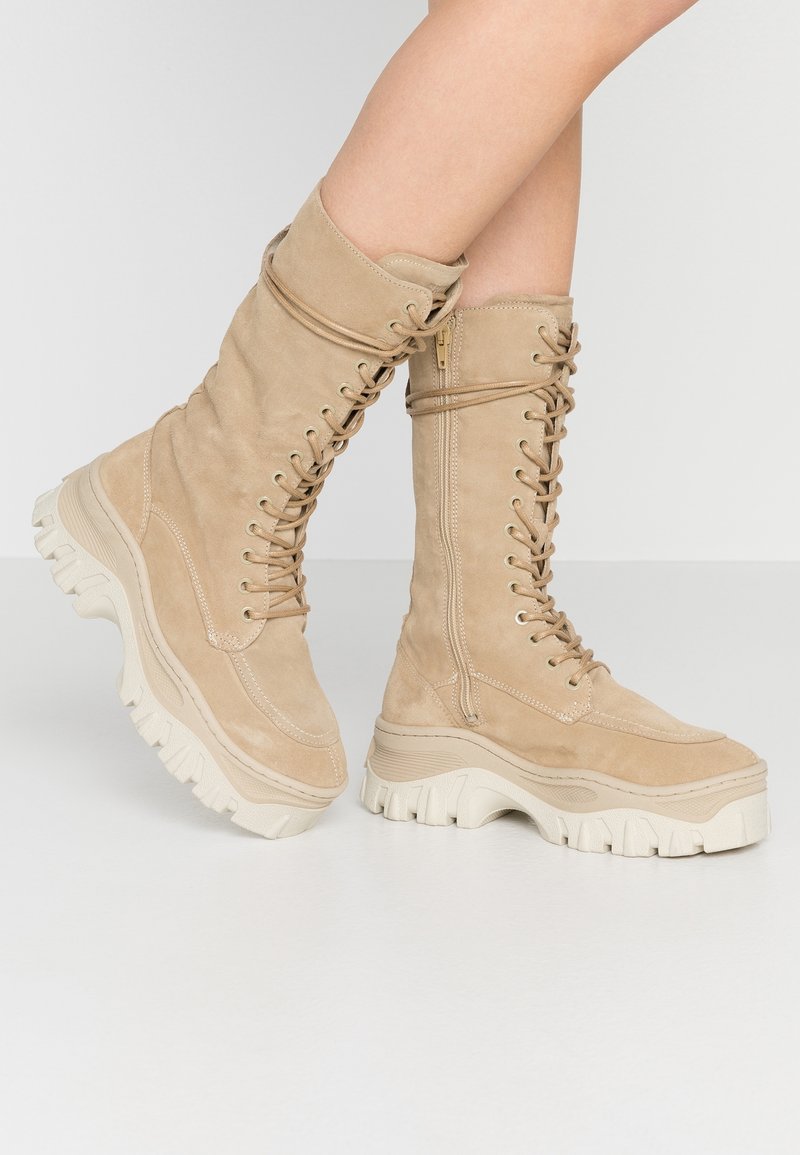 Botas de ante beige hasta la rodilla con cordones en el frontal, superficie texturizada, cremalleras en los laterales y suelas gruesas de color crema.