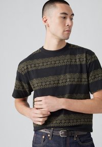 Camiseta negra de manga corta con rayas decorativas en verde oliva y un pequeño logo. Tejido de algodón con una textura suave.