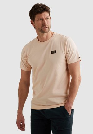 Muž v svetlobéžovom tričku s krátkym rukávom s logom na hrudi a v čiernych nohaviciach, stojaci s jednou rukou v vrecku, pozerajúci doľava.