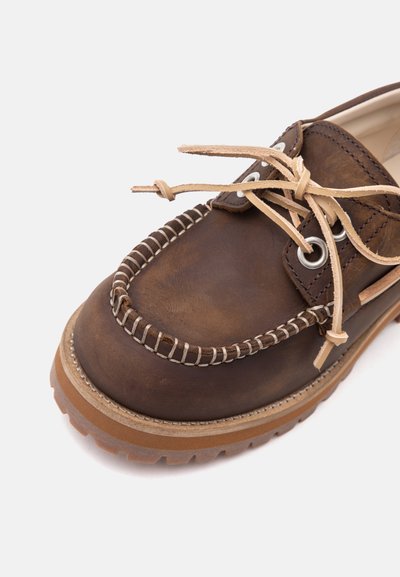 Mocassin en cuir marron avec des lacets beige noués en nœud, des coutures blanches le long du bout et une semelle en caoutchouc épaisse.